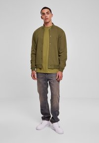 Mand der står iført en olivengrøn knapjakke, matchende grøn skjorte, falmede blå jeans og hvide sneakers foran en ensfarvet grå baggrund.