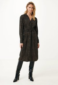 Femme debout dans une robe midi à manches longues, à motifs, avec une ceinture à la taille, assortie à des bottes à talons noires jusqu'aux genoux sur un fond uni.