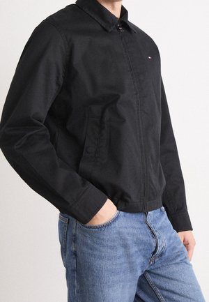 Mand iført en sort lynlåstrøje med krave og blå jeans, hånd stukket i jeanslommen, mod en ensfarvet lys baggrund.