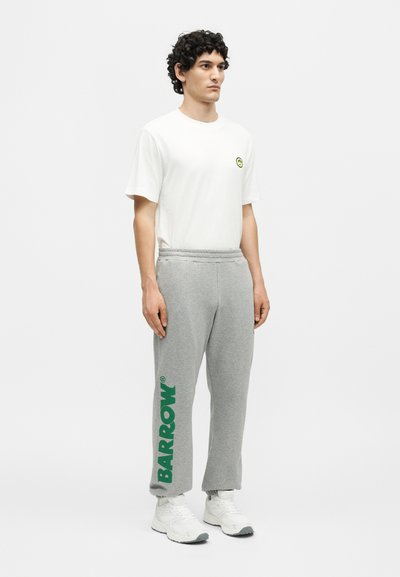 Άνδρας στέκεται φορώντας λευκό μπλουζάκι με μικρό κίτρινο σήμα χαμόγελου και γκρι παντελόνι φόρμας με μεγάλο πράσινο γράμμα "BARROW" στο αριστερό πόδι, λευκά αθλητικά παπούτσια.