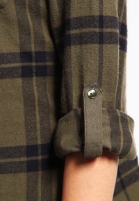 Grön rutmönstrad skjorta med knappar, tillverkad av mjukt tyg, med mörkblå rutig mönstring och upprullade ärmar med detaljer i metallknapp.