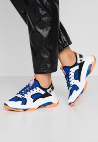 Scarpe atletiche con design bianco, blu e nero con accenti arancioni, mesh testurizzato e suola spessa per supporto.