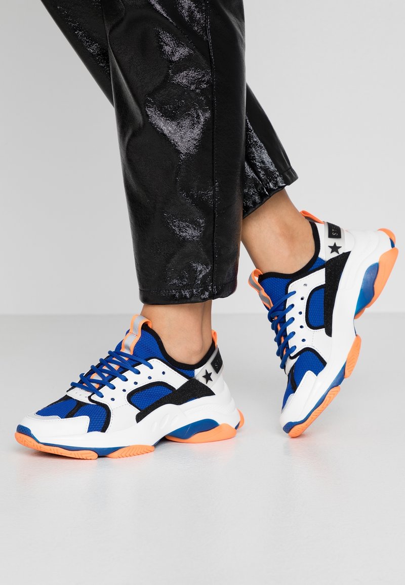 Scarpe atletiche con design bianco, blu e nero con accenti arancioni, mesh testurizzato e suola spessa per supporto.