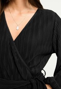 Zwarte geplooide wikkeljurk met een V-halslijn en een gestrikt taille. Beschikt over een delicate gouden ketting met een ovale hanger.