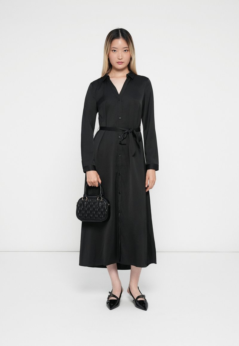 Zwarte jurk met lange mouwen, een knoopsluiting aan de voorkant en een strik in de taille, gecombineerd met een gestructureerde zwarte handtas en puntige zwarte ballerina's.