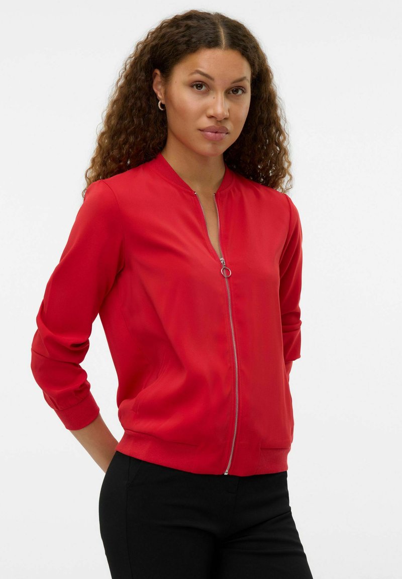 Kobieta o kręconych włosach, ubrana w czerwoną kurtkę na suwak z kółkiem zamiast suwaka i czarne spodnie, stoi z rękami za plecami.