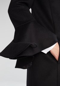 Sort frakke med flagrende, voluminøse ærmer. Har en glat tekstur, en skjult lomme og et kontrasterende hvidt indre lag, der er synligt ved manchetten.