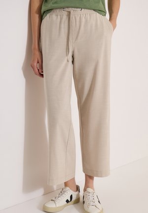 Personne portant un pantalon beige à jambes larges avec une taille élastique et des baskets blanches avec des détails noirs, debout contre un mur blanc.