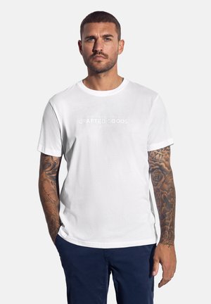 Hombre con el pelo corto y tatuajes en ambos brazos, viste una camiseta blanca de "Crafted Goods" y pantalones azul marino, de pie contra un fondo liso.