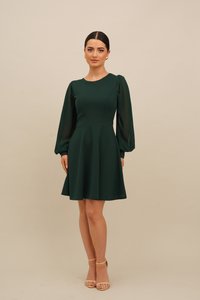 Mørkegrønn kjole med innsvingt liv og utsvingt skjørt. Har lange, gjennomsiktige ermer og rund halsringning. Brukt med nakne, reimede hæler.