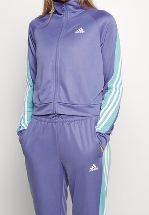 Person trägt violetten Adidas-Trainingsanzug mit hellblau-weiß gestreiften Ärmeln und Hosen, Hände in den Taschen, steht vor weißem Hintergrund.
