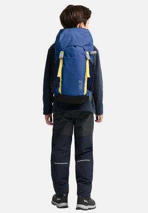 Młoda osoba zwrócona tyłem, nosząca niebiesko-żółty plecak, ciemne ubrania outdoorowe oraz czarne buty trekkingowe na jednolitym tle.