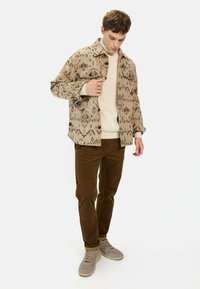 Beige geblokte shirtjack met geometrische prints, crèmekleurige trui, bruine corduroypantalon en grijze sneakers met lichte accenten.