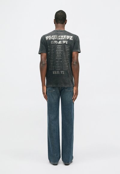 Hombre de espaldas, vistiendo una camiseta de banda gráfica negra y jeans azules sueltos, apoyado contra un fondo blanco liso.