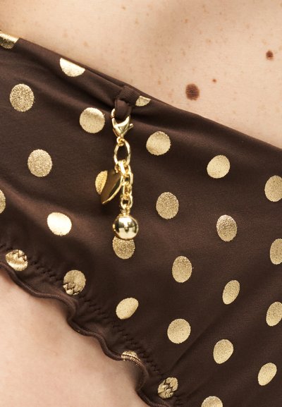 Close-up of brown fabric with gold polka dots and a small gold charm hanging from a fabric loop against light skin with freckles and moles.