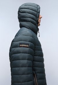 Giubbotto imbottito in verde teal scuro, con cappuccio, texture trapuntata, patch del logo sulla manica sinistra e dettagli con zip laterali.