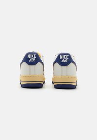 Une paire de baskets Nike Air blanches avec un onglet de talon bleu et une semelle intermédiaire beige, vues de l'arrière sur un fond blanc.