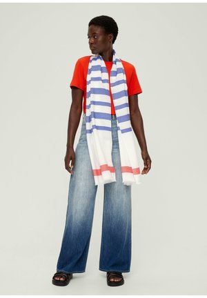 Écharpe blanche avec des rayures bleues et rouges, portée par-dessus une chemise orange. Modèle en jean large et sandales noires, se tenant devant un fond neutre.
