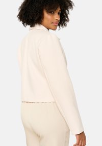 Gai bežs īsais žakets ar teksturētu apdari, ar iedobtu apkakli un garām piedurknēm, attēlots no aizmugures.