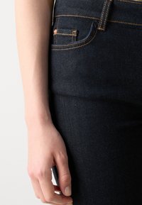 Mörkblå denimjeans med orange sömmar, med en framficka och en slät textur. Hand vilande på midjebandet.