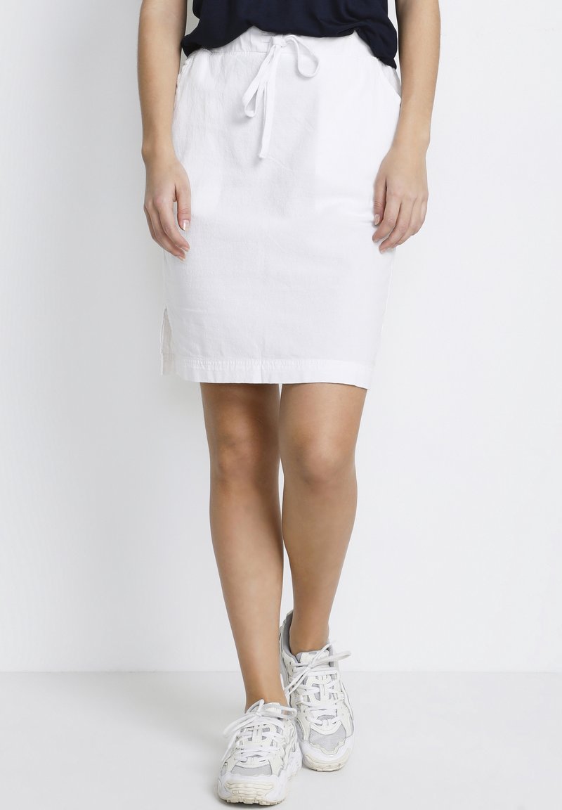 Vit linnekjol med dragsko i midjan, medellång, med sidoklappar för bättre rörelsefrihet. Kombinerad med grå och vita sneakers.