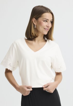 Woman with shoulder-length brown hair wearing a white V-neck blouse with lace-trimmed sleeves and black pants, smiling and looking to the side.