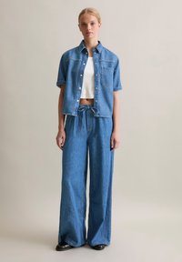 Denim shirt with short sleeves and a pocket, paired with wide-leg jeans, both in medium blue. White ribbed crop top underneath. Black shoes.