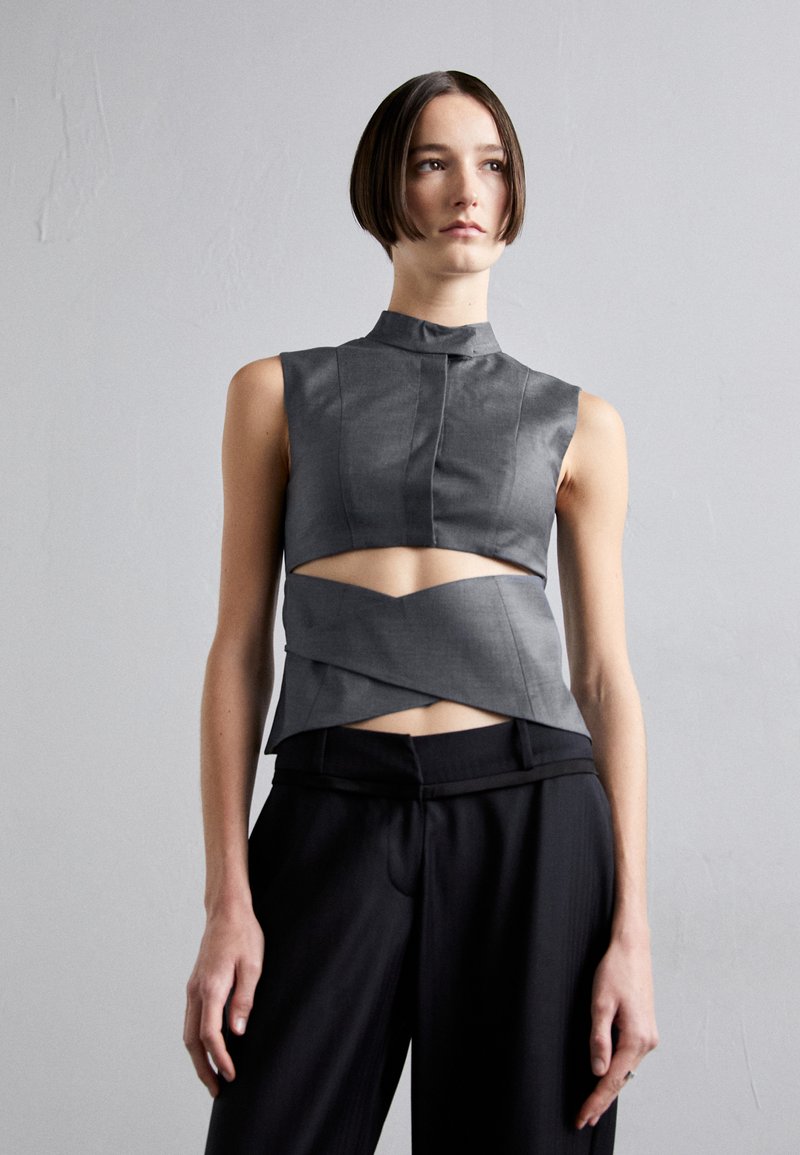 Gray sleeveless top with a high collar, featuring cut-out details and a layered design. Paired with black tailored trousers.
