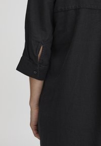 Chemise noire texturée avec un poignet à bouton et un détail fente. La vue arrière montre une coupe décontractée et un tissu doux.