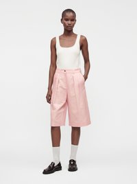 Mannequin mince portant un haut blanc sans manches, un short rose taille haute arrivant aux genoux, des chaussettes blanches et des mocassins noirs, debout devant un fond uni.