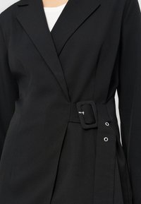 Gros plan sur un blazer noir avec un large col et une ceinture en tissu dotée d'une boucle rectangulaire et d'œillets métalliques sur le côté.