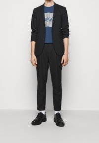 Czarna marynarka na niebieskiej koszulce z szarym logo, w zestawieniu z czarnymi spodniami i czarnymi sznurowanymi butami. Widoczne białe skarpetki. Gładkie faktury.