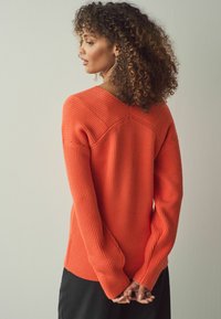 En orange ribbad tröja med V-ringning och långa ärmar, med en avslappnad passform och texturerad tyg, sedd bakifrån.