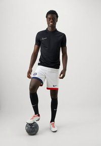 Roupa desportiva Nike com uma camisa preta de manga curta, calções brancos com detalhes em azul e vermelho, meias pretas até ao joelho e chuteiras brancas.