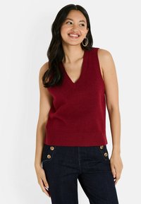 Femme souriante avec de longs cheveux noirs portant un pull sans manches en tricot bordeaux et un jean foncé avec des boutons en bois sur les poches.