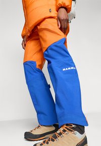 Pantaloni da outdoor Mammut con blocchi di colore arancione e blu, tessuto testurizzato, zip laterali e rinforzi nelle ginocchia, abbinati a scarponi da trekking beige.