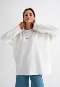Witte oversized sweatshirt van zacht materiaal, met een ronde hals en het woord "MIRONS" in bruin geborduurd op de borst.