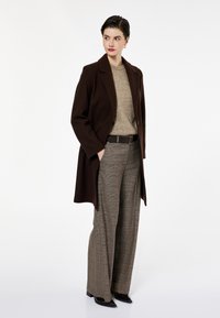 Ruskotoinen villakangastakki, joka on muotoiltu istuvaksi, vaaleanbeige villapaita sen alla ja ruudulliset housut. Mustat korot täydentävät asun. Yksinkertainen tausta.