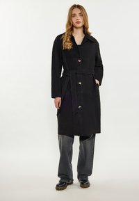 Černý trench coat s páskem a límcem, s kovovými knoflíky, bočními kapsami a hladkým povrchem, doplněný šedými širokými kalhotami.