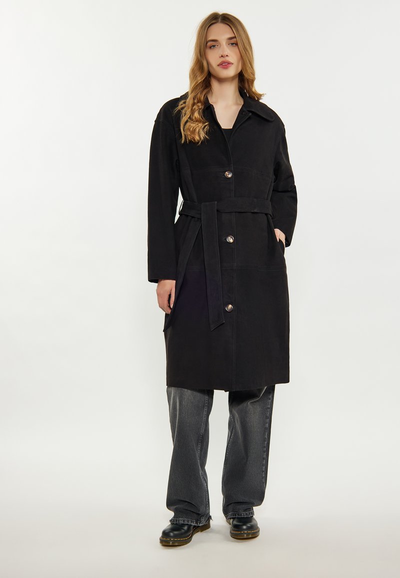 Černý trench coat s páskem a límcem, s kovovými knoflíky, bočními kapsami a hladkým povrchem, doplněný šedými širokými kalhotami.