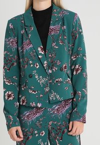 Veston bleu sarcelle avec un motif floral et plumes, design croisé, boutons noirs et manches longues, assorti à un pantalon assorti.