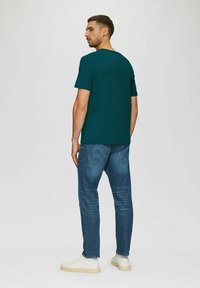 Türkisfarbenes T-Shirt mit kurzen Ärmeln und rundem Ausschnitt, kombiniert mit hellblauen schmalen Jeans und weißen Sneakers, von hinten gezeigt.
