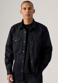 Black denim shirt with silver snap buttons, two chest pockets, and a collar. Worn over a black T-shirt, featuring a classic fit.