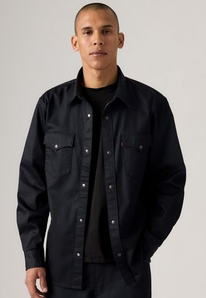 Black denim shirt with silver snap buttons, two chest pockets, and a collar. Worn over a black T-shirt, featuring a classic fit.