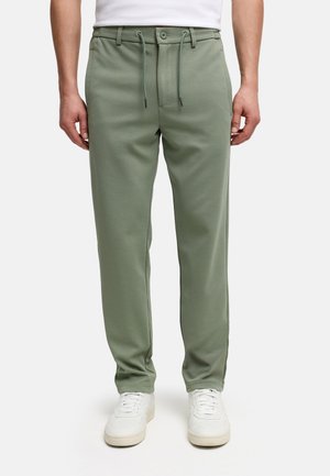 Hombre que lleva pantalones casuales verdes con textura, cintura con cordón y zapatillas blancas, de pie frente a un fondo liso.