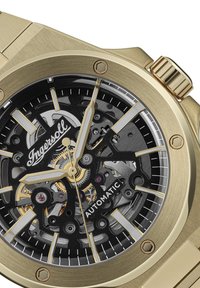 Goudkleurig automatisch horloge met een transparante zwarte skeleton-dial, gouden accenten en luminescente uurmarkeringen. Voegt industriële details toe met een metalen textuur.