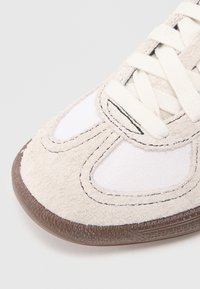 Sneaker, amelynek fehér vászon felsőrésze bézs velúr részletekkel, kerek orral és texturált barna gumitalppal rendelkezik. A fűző krémszínű.