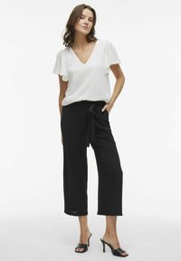 White V-neck blouse with ruffled sleeves, paired with black wide-leg trousers featuring a tie waist and smooth texture, complemented by black heels.
