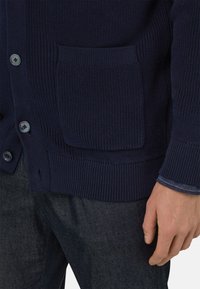 Cardigan in maglia blu navy con sei bottoni, texture a coste e dettaglio con tasca frontale. Indossato sopra una camicia blu con bottoni e pantaloni in denim.