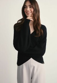 Młoda kobieta z długimi brązowymi włosami, uśmiechająca się, ubrana w czarny luźny sweter i jasne szerokie spodnie na jednolitym tle.